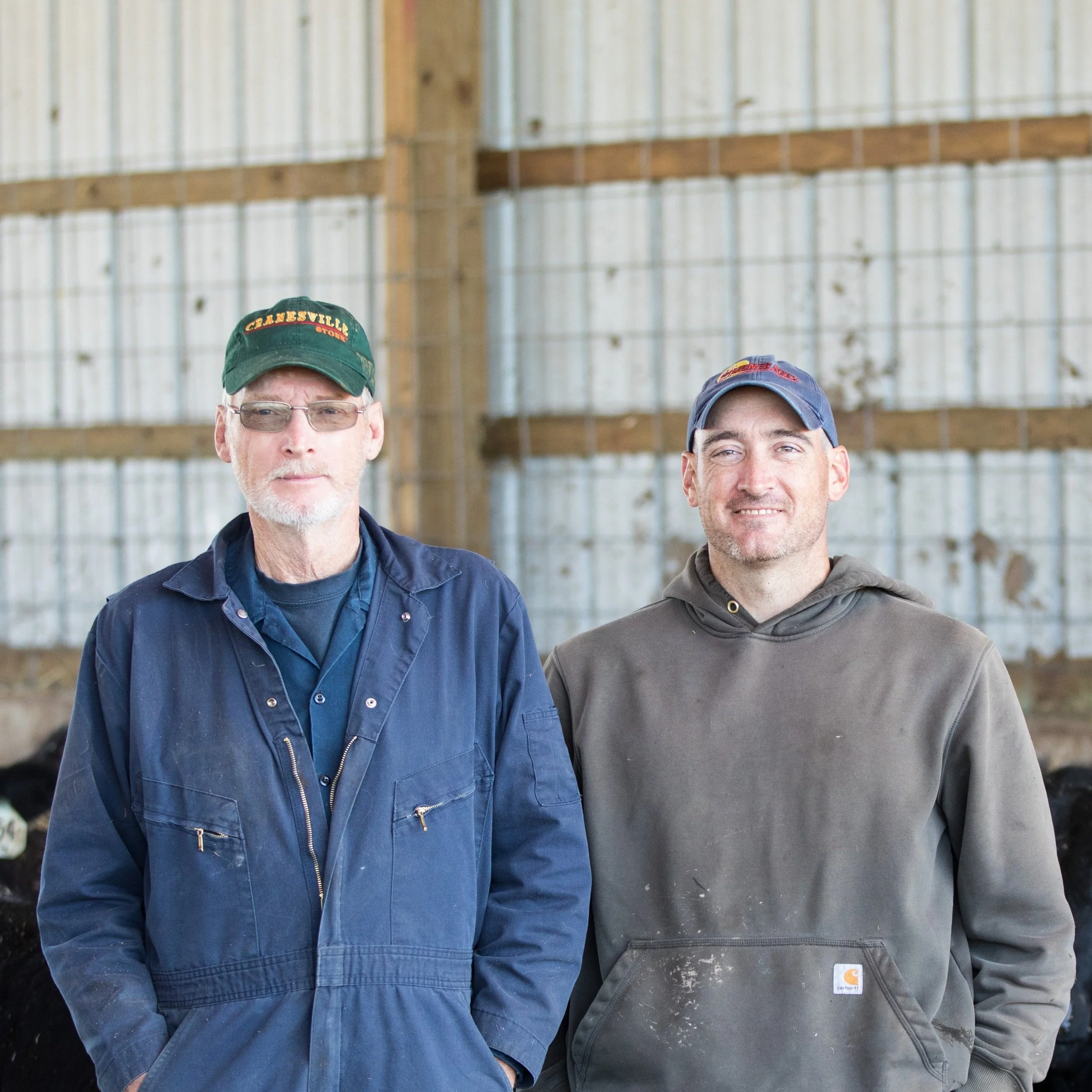 Two farmers standing together in a barn, wearing work clothes and hats, with a rustic wooden background. The image captures their connection to agriculture and livestock farming.